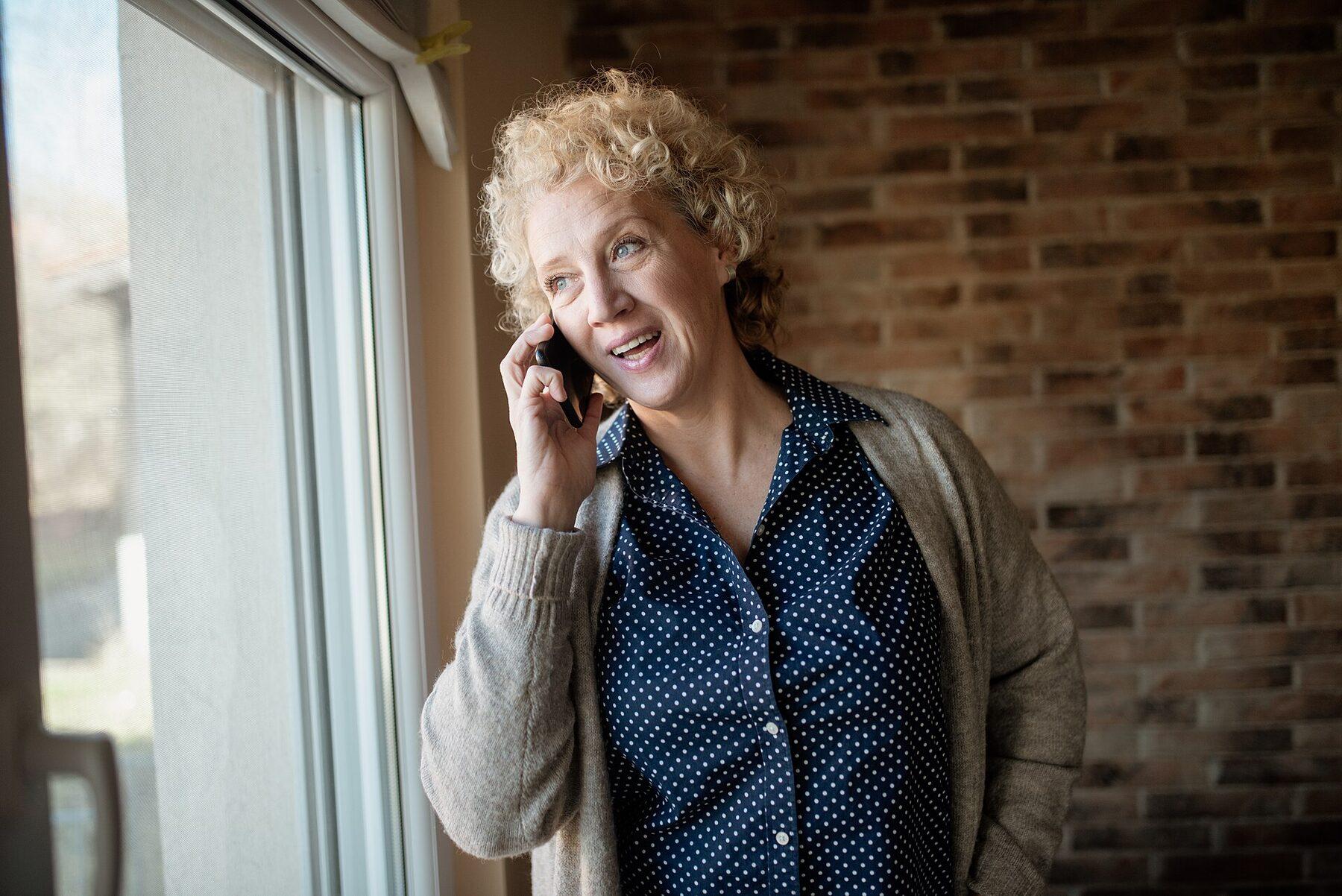 A woman smiles while talking on her phone beside a window.